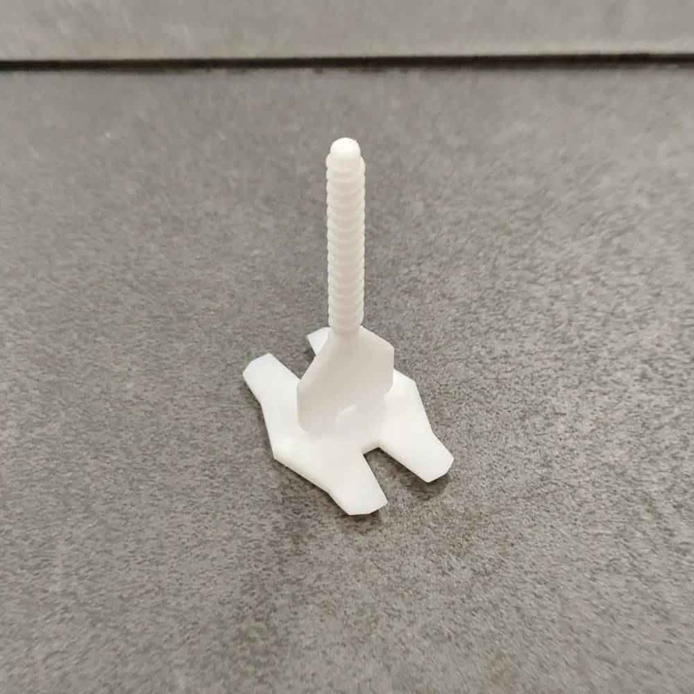 Eine kleine, weiße Kunststoffschraube mit einer flachen Basis steht aufrecht auf einer strukturierten grauen Oberfläche.