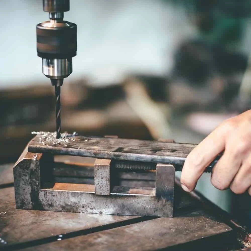 Eine Hand benutzt eine Ständerbohrmaschine, um ein Loch in ein Metallstück zu bohren, das von einem Schraubstock gehalten wird, was einen Metallbearbeitungsprozess in einer Werkstattsituation darstellt.