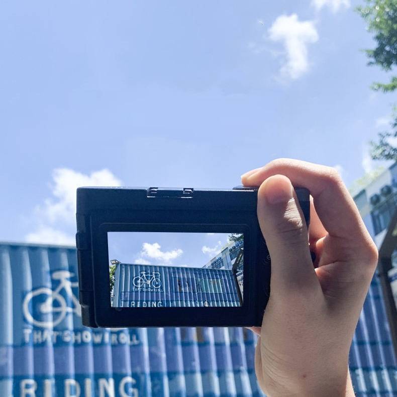 Eine Hand hält ein Smartphone und fotografiert ein blaues Gebäude mit einem Fahrradbild und dem teilweisen Text „RIDING