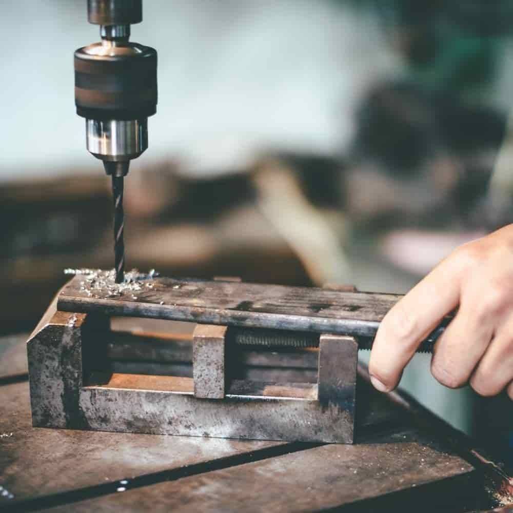 Eine Person bedient eine Bohrmaschine in einer Werkstatt und konzentriert sich auf präzises Bohren in ein Metallteil, das in einem Schraubstock befestigt ist.