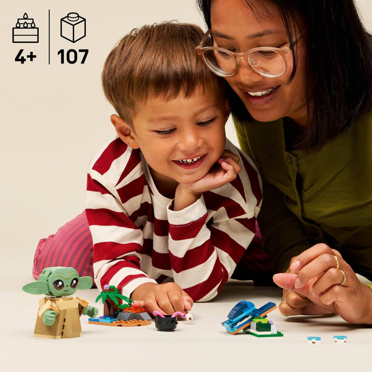 Ein kleines Kind und ein Erwachsener spielen mit LEGO-Steinen und bauen eine kleine Szene mit verschiedenen Strukturen und einer grünen Figur. Der Text lautet „4+ 107