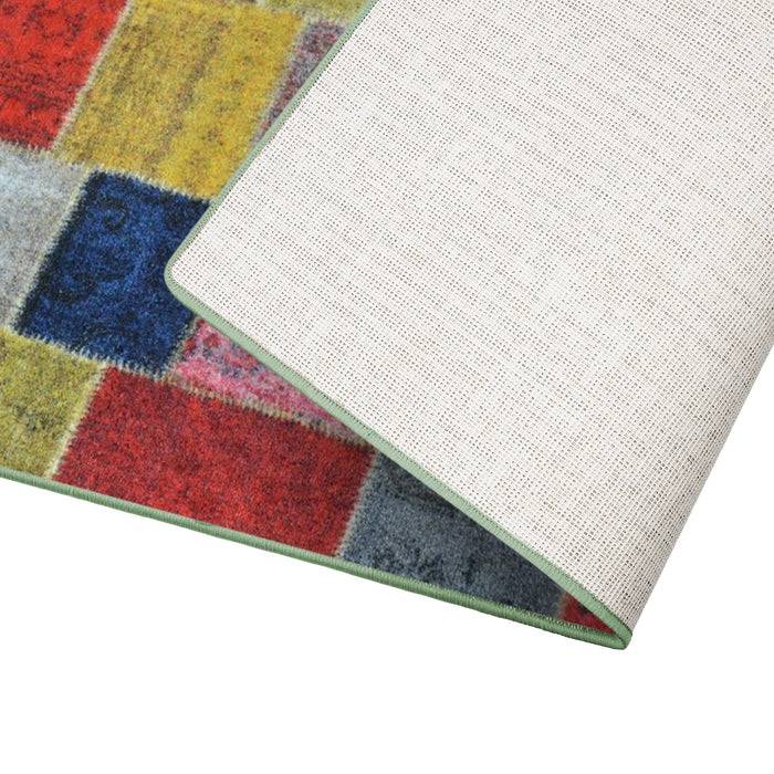 Ein farbenfroher gewebter Teppich mit roten, blauen, gelben und grauen Quadraten ist teilweise gefaltet, um seine Stoffstruktur und grüngefasste Kante zu zeigen.