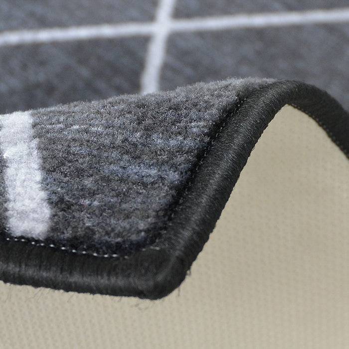 Nahaufnahme eines grauen Teppichs mit schwarzer Borte, die seine strukturierte Webung und rutschfeste Unterseite zeigt, auf einem gefliesten Boden platziert.