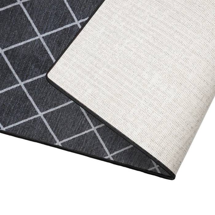 Ein schwarzer Teppich mit einem weißen geometrischen Kreuzmuster, teilweise aufgerollt dargestellt, wobei seine helle Unterseite und schwarze Kante sichtbar sind.