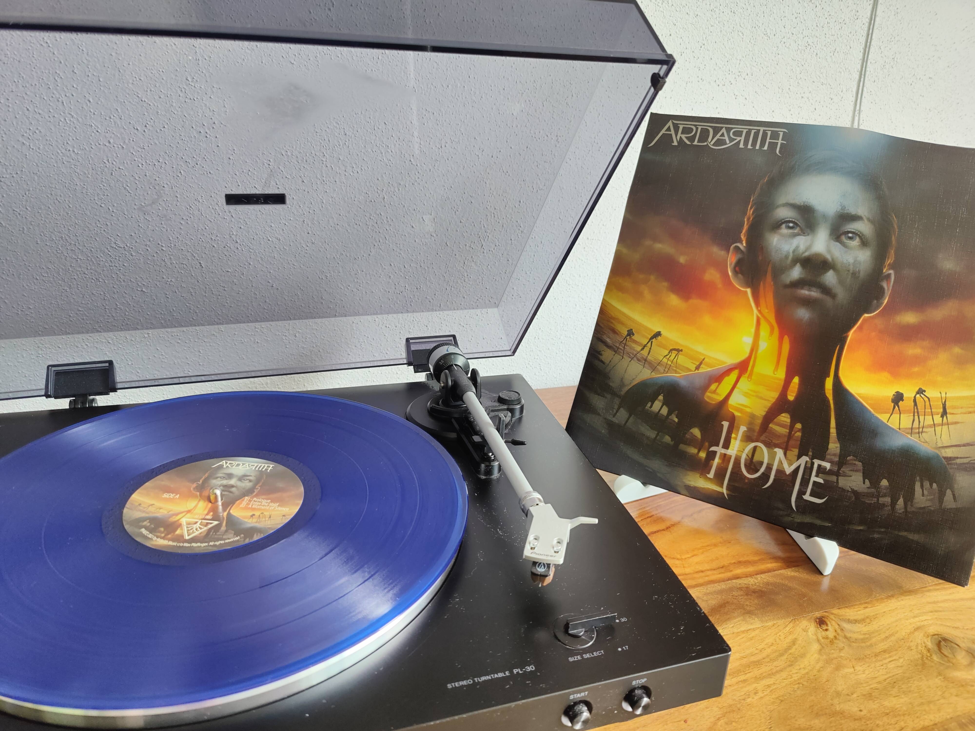 Ein schwarzer Plattenspieler mit einer blauen Schallplatte beim Abspielen. Daneben zeigt ein Albumcover ein Porträt einer Person mit industriellen Elementen im Hintergrund.