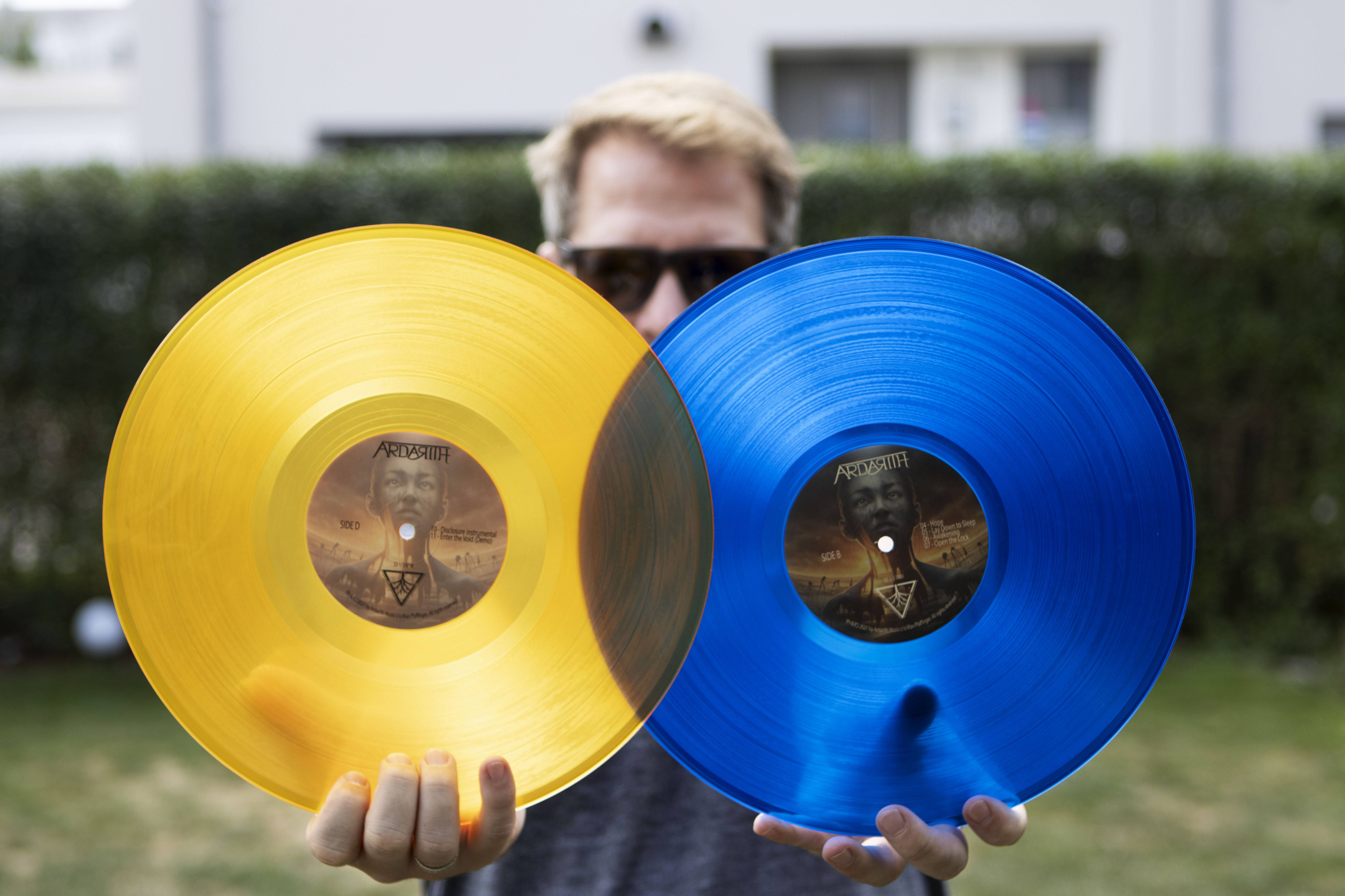 Eine Person hält zwei durchscheinende Schallplatten aus Vinyl, eine gelbe und eine blaue, jede mit einem Etikett, das ein dreieckiges Logo und Text zeigt.