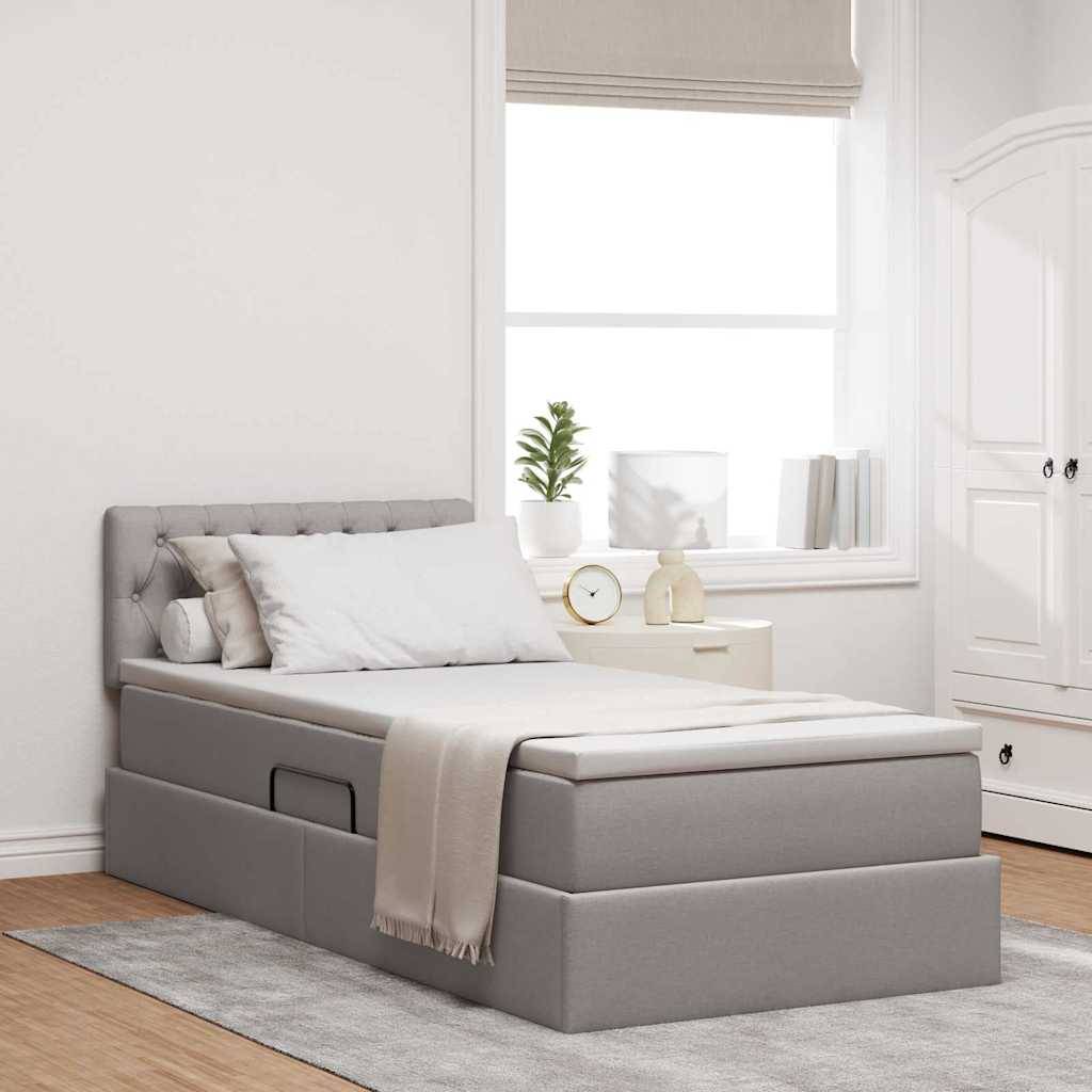 Ein modernes Schlafzimmer mit einem grau gepolsterten Bett mit Schubladen zur Aufbewahrung, einer beige Wurfdecke und einem Nachttisch mit Dekorationsgegenständen neben einem sonnigen Fenster.