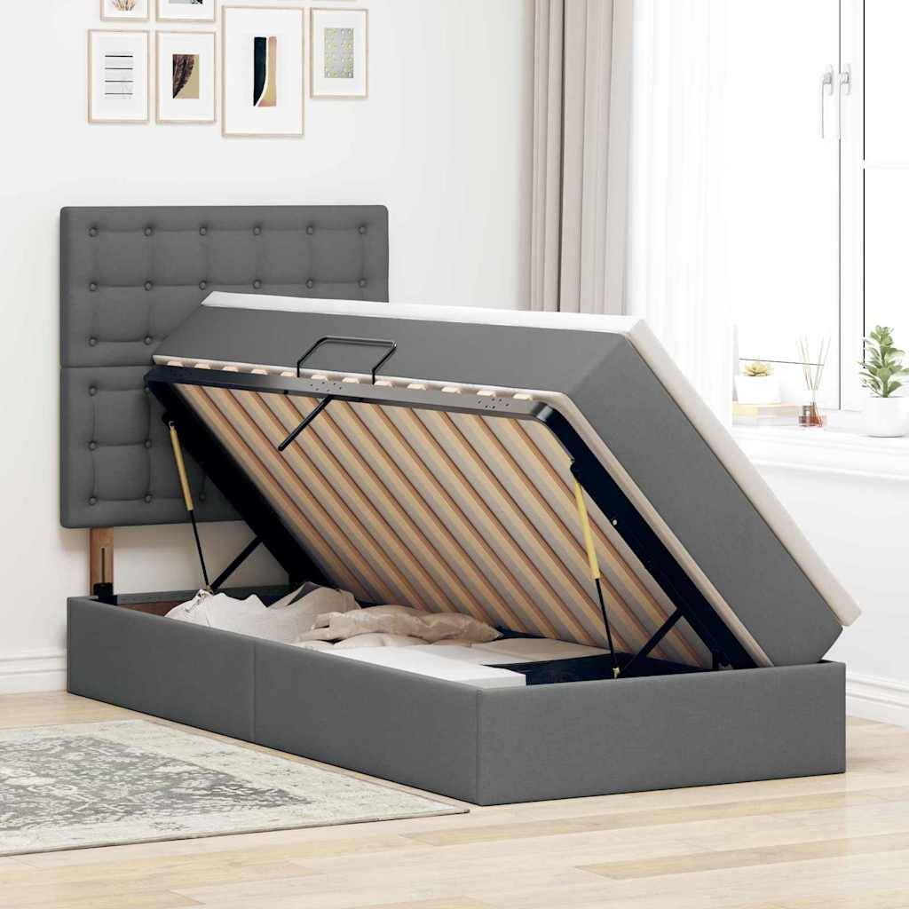 Ein grau gepolstertes Speicherbett mit offener hydraulischer Hubbühne zeigt innen aufbewahrte Decken. Es steht in einem hellen Zimmer mit gerahmter Kunst und einem Fenster.