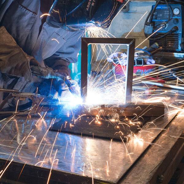 Ein Schweißer in Schutzausrüstung und Maske schweißt einen Metallrahmen auf einer Werkbank, wobei helle Funken in der gesamten Werkstatt versprüht werden.