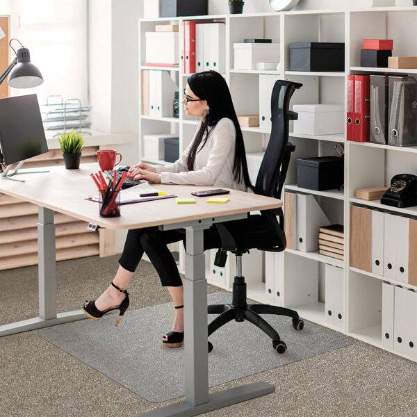 Frau, die an einem Schreibtisch in einem hellen Büro arbeitet, sitzt auf einem schwarzen Stuhl neben einem Computer und Regalen mit Ordnern.