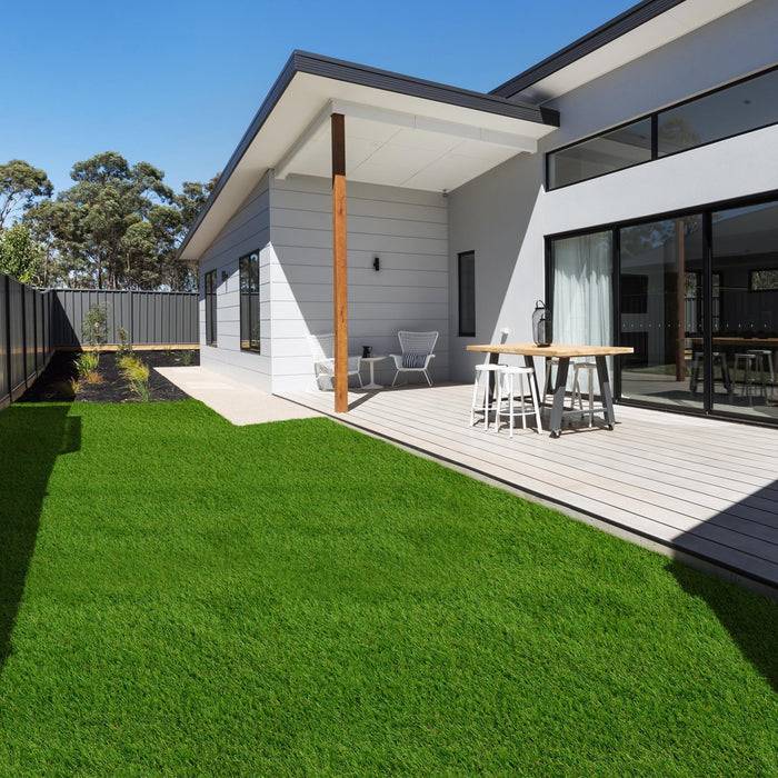 Modernes Haus mit großen Fenstern, einer Holzterrasse mit Außensitzplätzen und einem ordentlich gestutzten grünen Rasen, der von einem Zaun begrenzt wird.