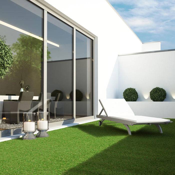Eine moderne Terrasse mit grünem Rasen, einem weißen Liegestuhl und einem Glastisch mit Stühlen. Große Glastüren führen ins Haus.