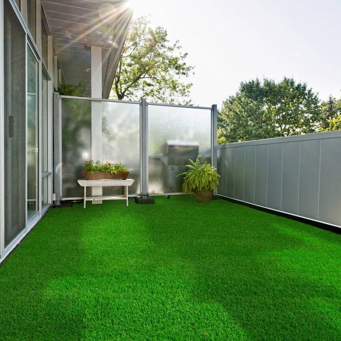 Ein sonniger Balkon mit grünem Kunstrasen, Topfpflanzen und Blick auf Bäume, umrahmt von einem modernen Metall- und Glasgeländer.
