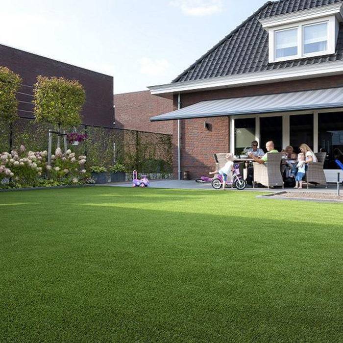 Eine Familie sitzt an einem Tisch auf einer Terrasse, die teilweise von einem Sonnenschirm bedeckt ist, neben einem Haus mit einem Garten und einem gepflegten Rasen.