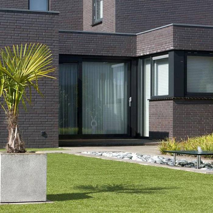 Ein modernes Backsteinhaus mit großen Glastüren, eine kleine Palme in einem quadratischen weißen Blumenkübel und ein ordentlicher grüner Rasen im Vordergrund.