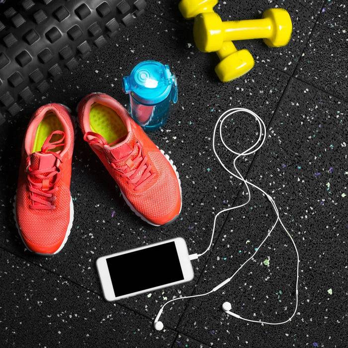 Rote Sportschuhe, eine blaue Wasserflasche, gelbe Hanteln, ein Smartphone und Kopfhörer liegen auf einem schwarz gesprenkelten Fitnessstudio-Boden.