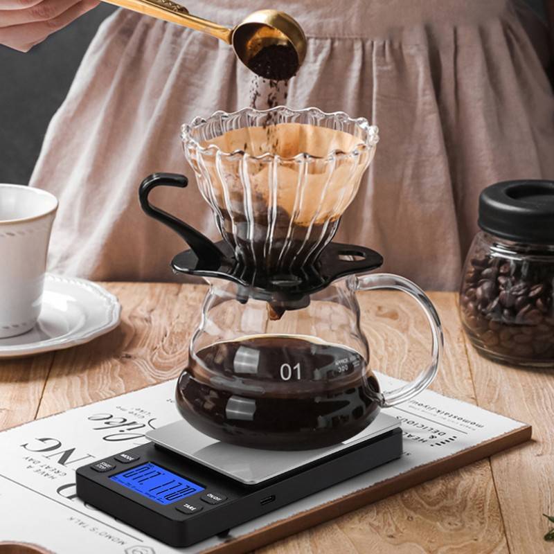 Kaffeepulver wird in einen Filter auf einer Glaskanne auf einer digitalen Waage gegossen, mit einer Kaffeetasse und einem Glas mit Kaffeebohnen in der Nähe, auf einem Holztisch.
