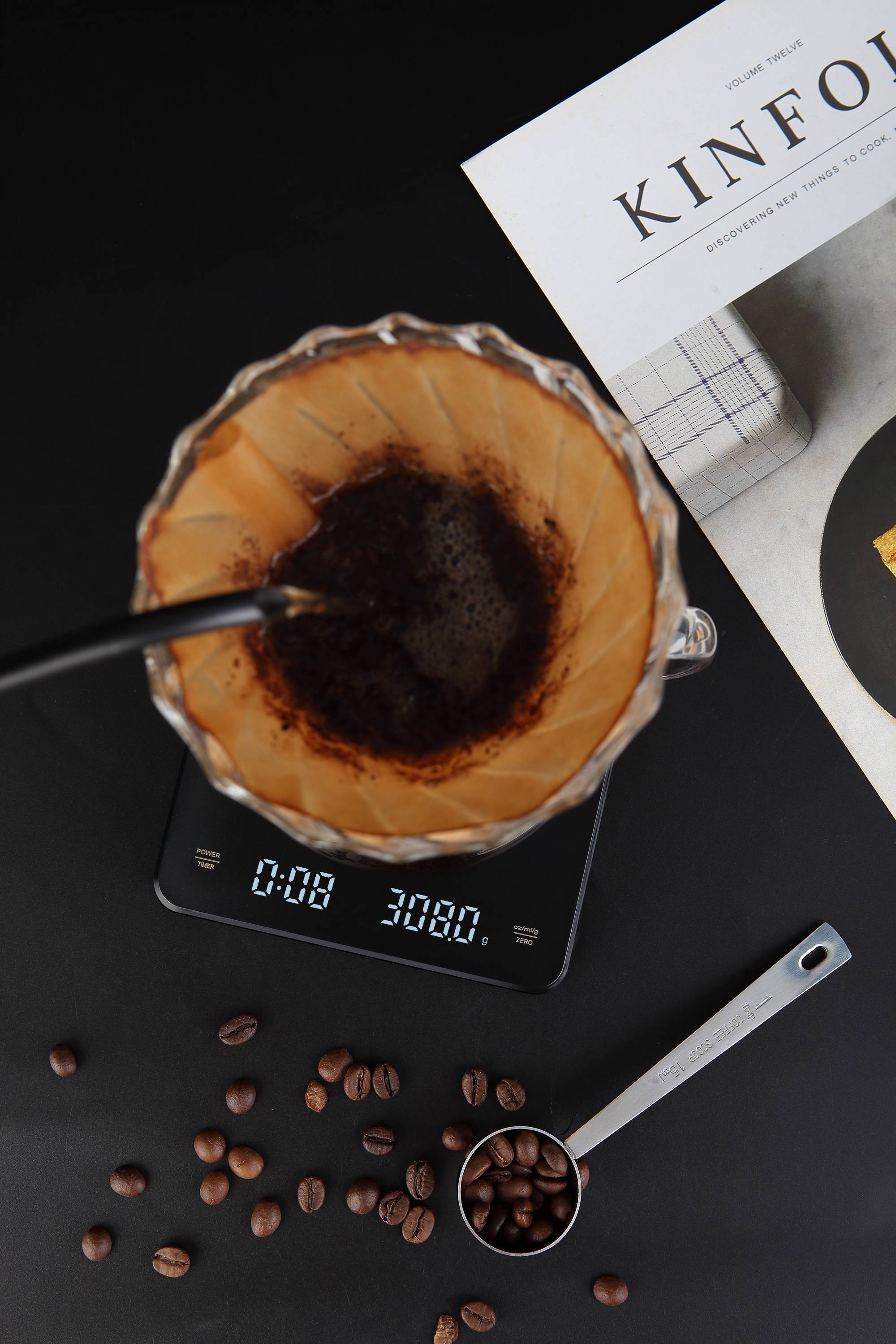 Aufsicht auf die Kaffeezubereitung mit der Pour-over-Methode. Ein Filter sitzt auf einer Waage, mit Bohnen daneben verstreut und ein Notizbuch teilweise sichtbar.