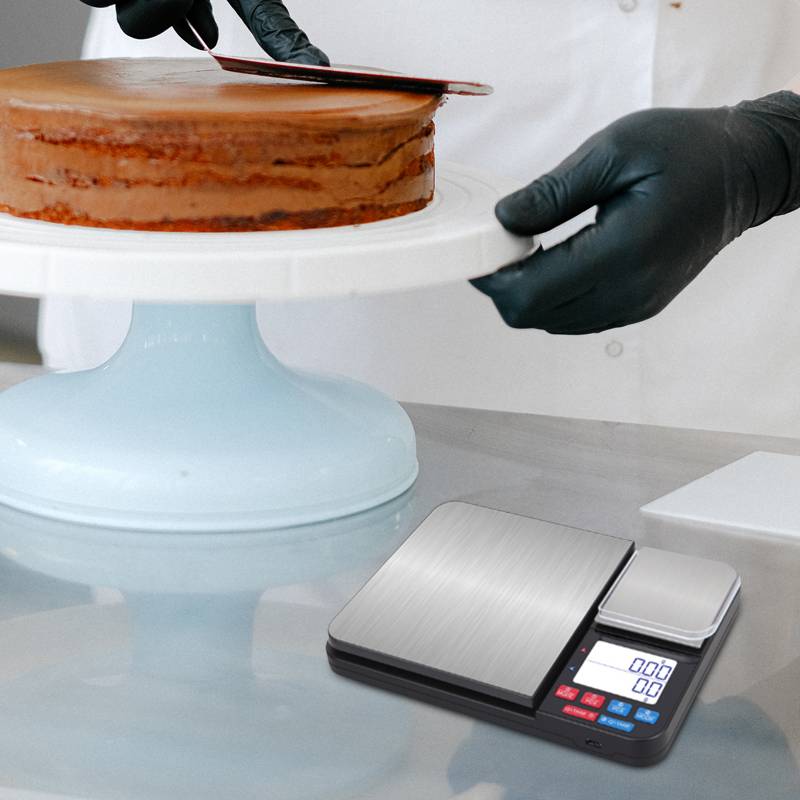 Eine Person mit Handschuhen glättet Schokoladenglasur mit einem Spachtel auf einem Kuchen. Eine elektronische Waage steht in der Nähe auf dem Tisch.