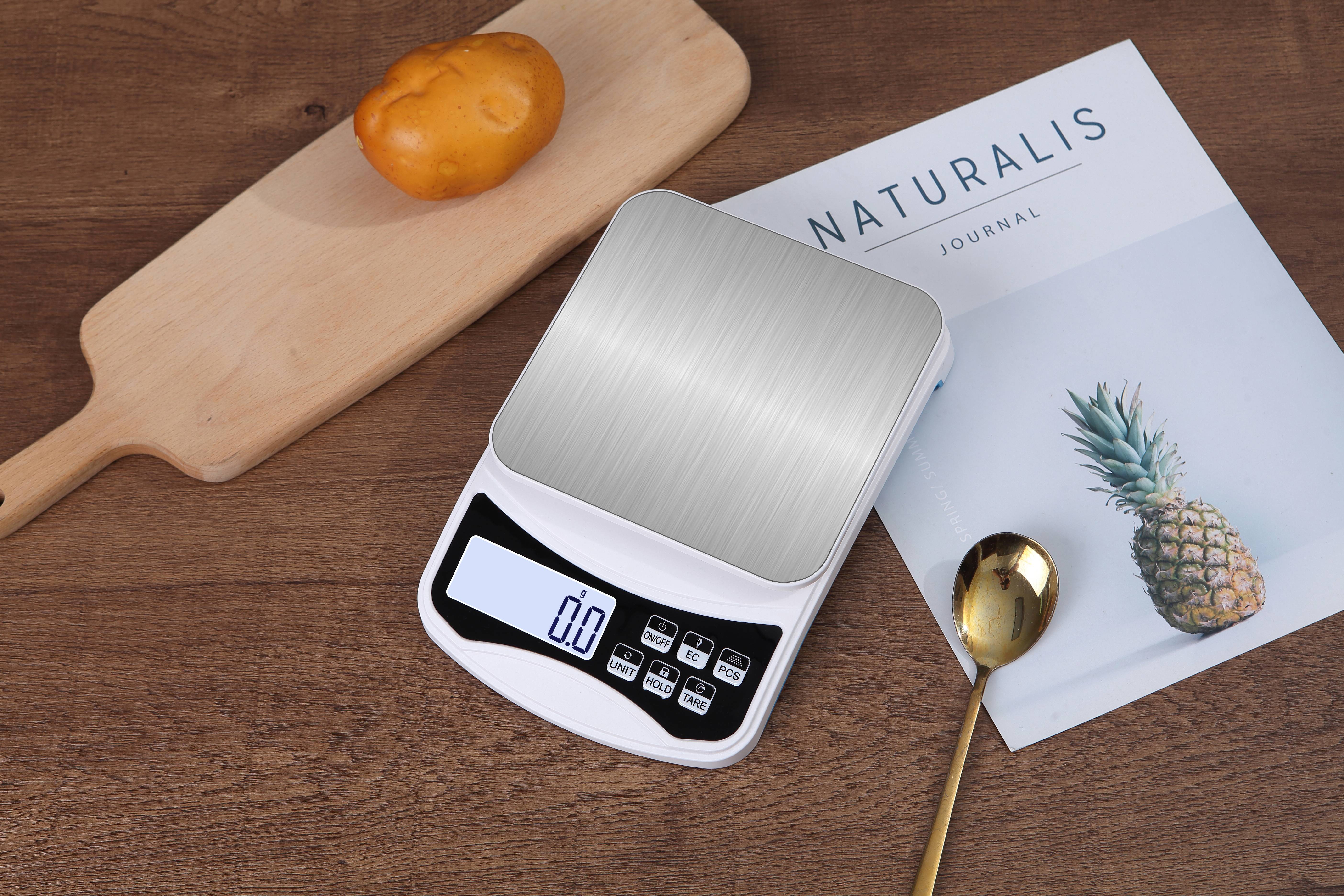 Digitale Küchenwaage zeigt 0,0 an, daneben ein goldener Löffel und ein Brötchen auf einem Holztisch, neben einem Journal mit dem Titel 