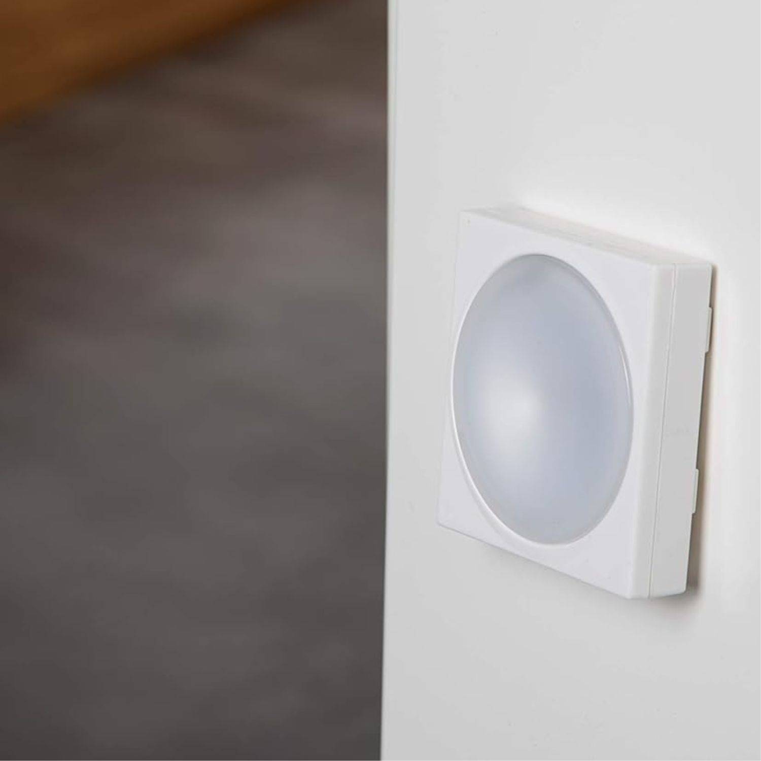 Ein runder, weißer Bewegungsmelder ist an einer weißen Wand montiert, ragt leicht hervor und hat einen sanften Glanz an seinen Kanten, was darauf hindeutet, dass er aktiv ist.