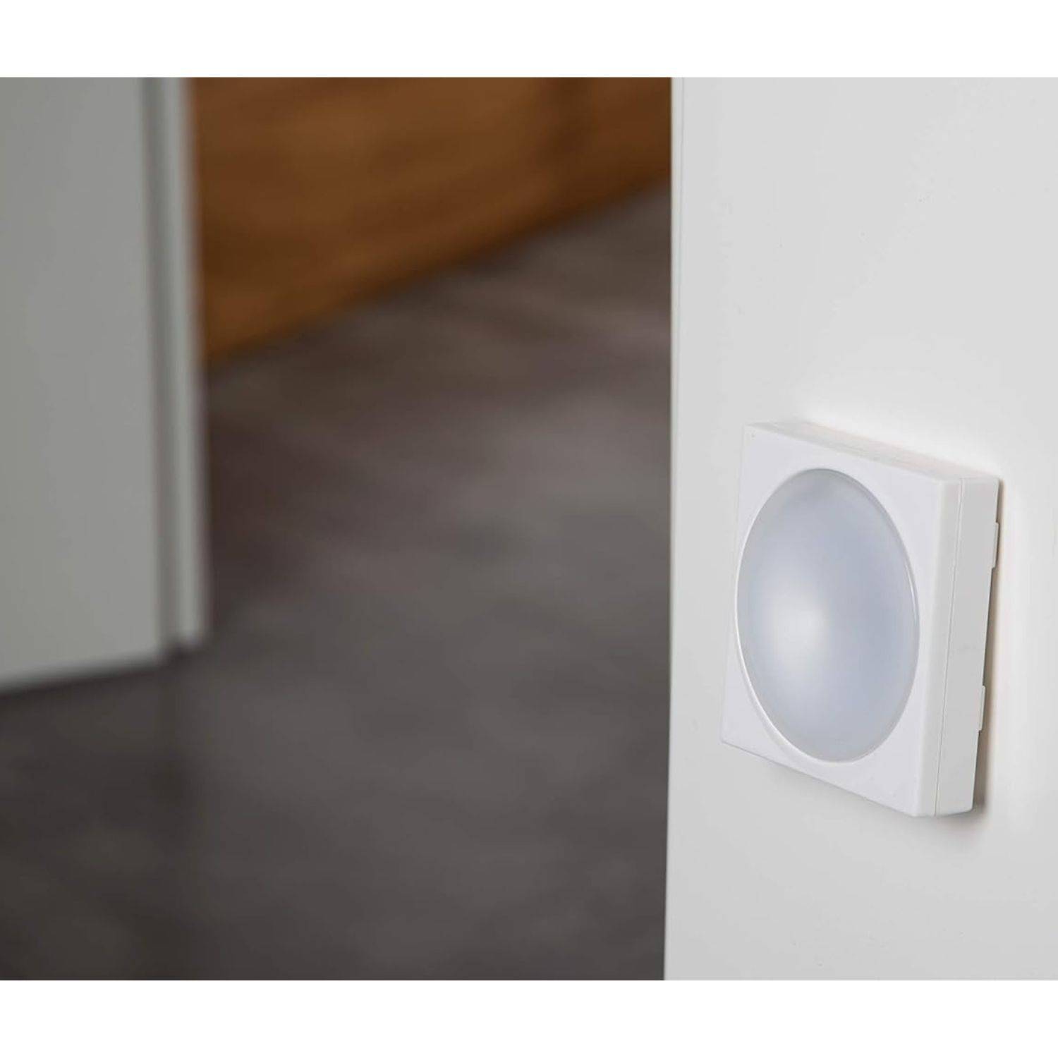 Eine an der Wand montierte weiße Wandleuchte mit rundem, mattem Diffusor, angebracht an einer weißen Wand. Eine Türöffnung mit unscharfem Innenraum ist sichtbar.