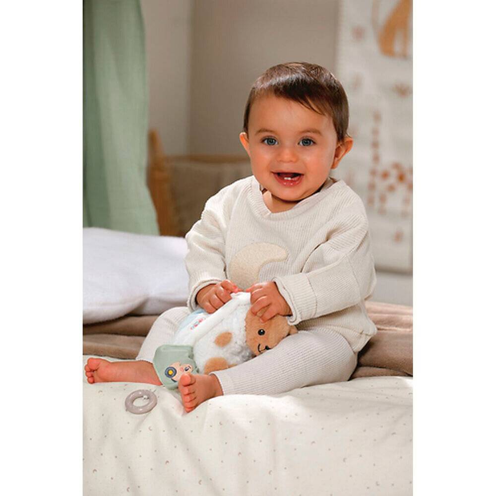 Ein lächelndes Baby trägt einen leichten Pullover und eine Hose und sitzt auf einem Bett, während es ein weiches, tierförmiges Spielzeug hält. Das Zimmer ist sanft beleuchtet und gemütlich.