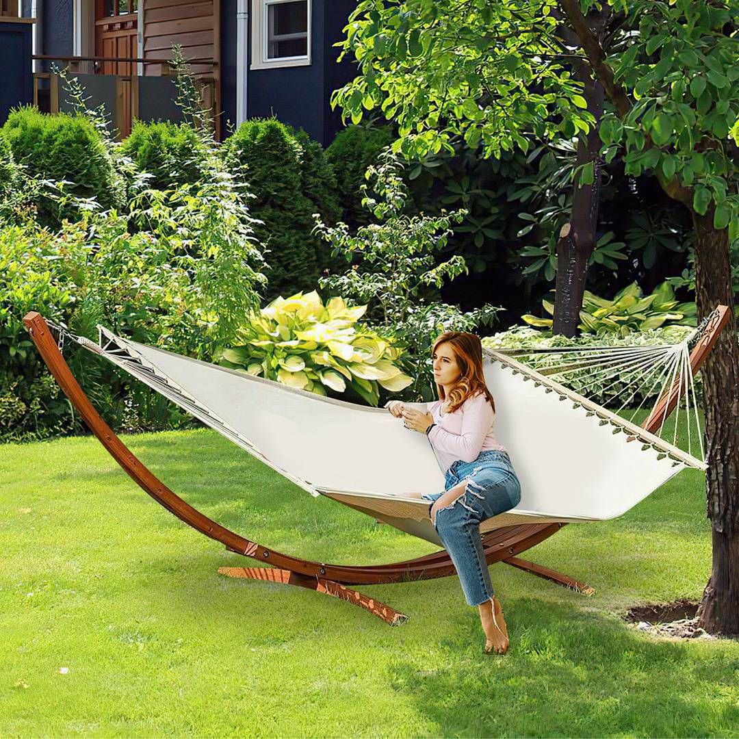 Eine Frau sitzt in einer Hängematte in einem üppigen Garten, umgeben von grünen Sträuchern und Bäumen, mit einem Haus, das im Hintergrund sichtbar ist.