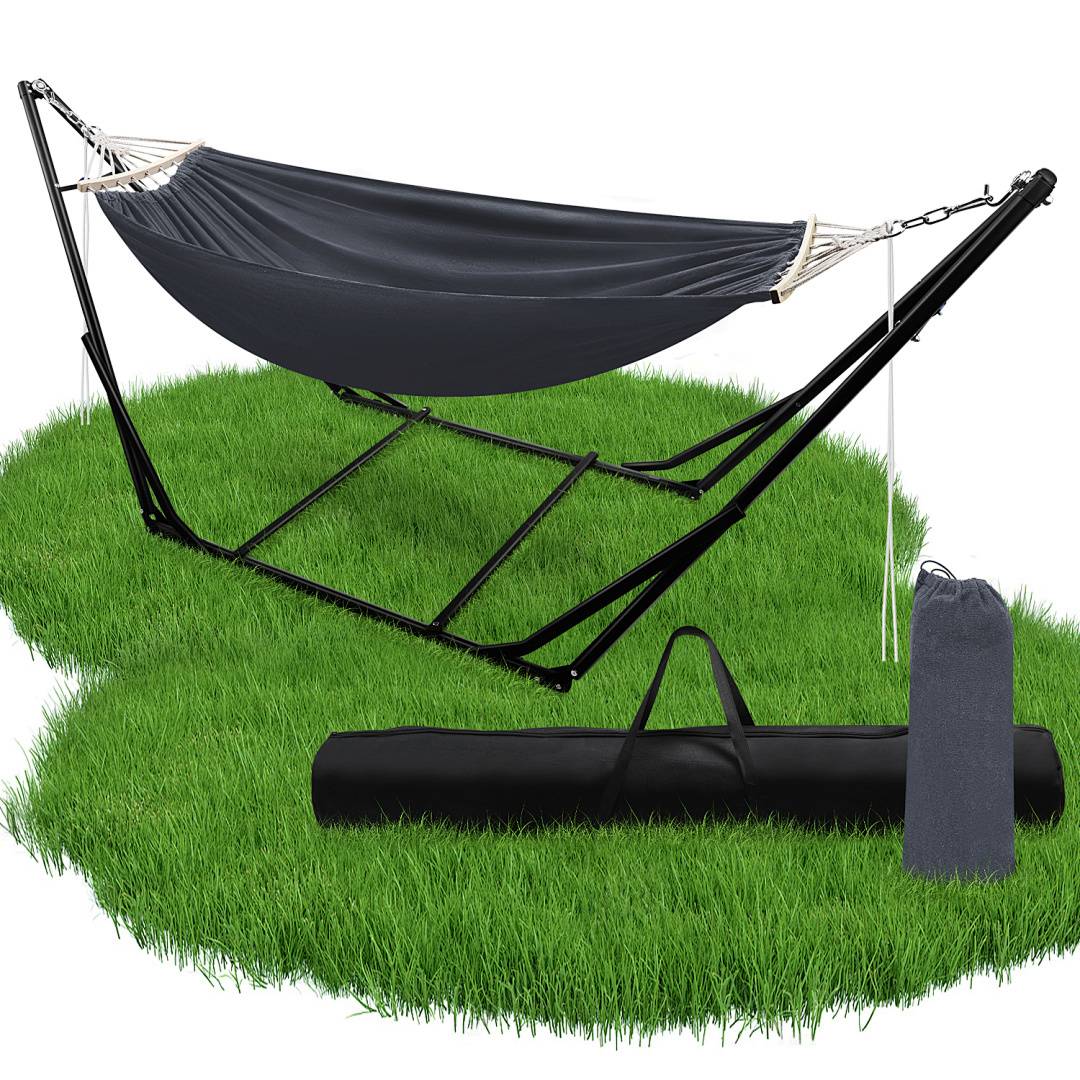 Eine schwarze Stoff-Hängematte ist an einem Metallgestell aufgehängt, das auf Gras steht. Eine Tragetasche und ein grauer Stoff-Beutel sind neben dem Aufbau platziert.