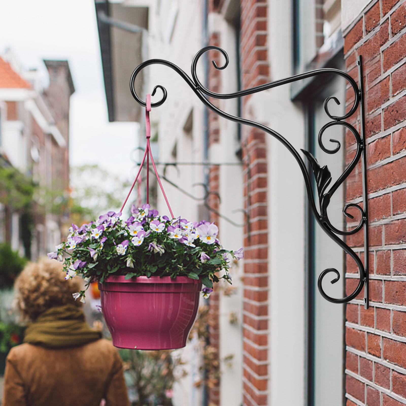 Ein rosa Blumentopf mit blühenden violetten und weißen Blumen hängt an einer kunstvollen Metallhalterung an einer Backsteinmauer. Eine Person geht vorbei.