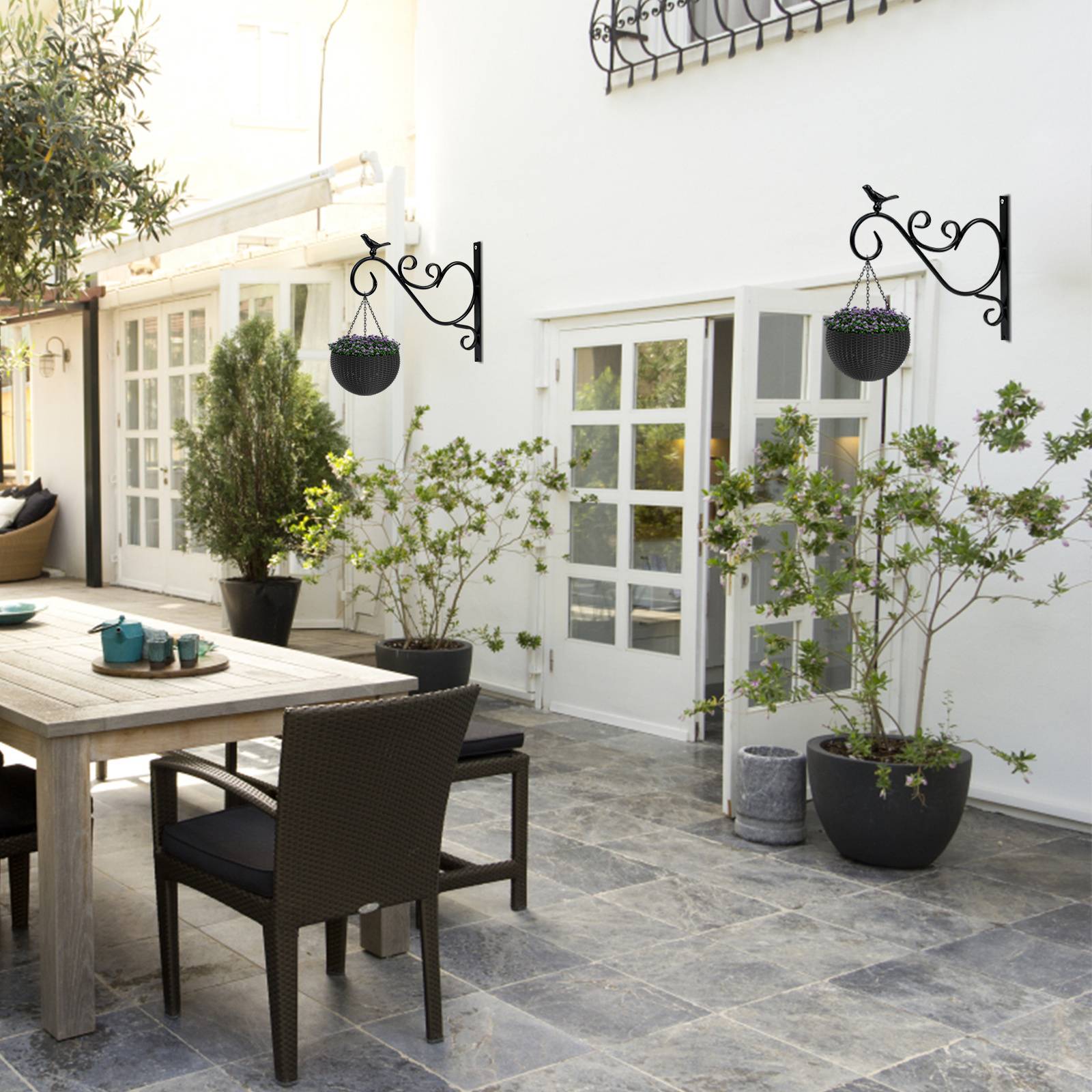 Eine gemütliche Außenterrasse mit einem Holztisch, schwarzen Korbstühlen, Topfpflanzen und dekorativen Wandleuchten an einem weißen Gebäude.