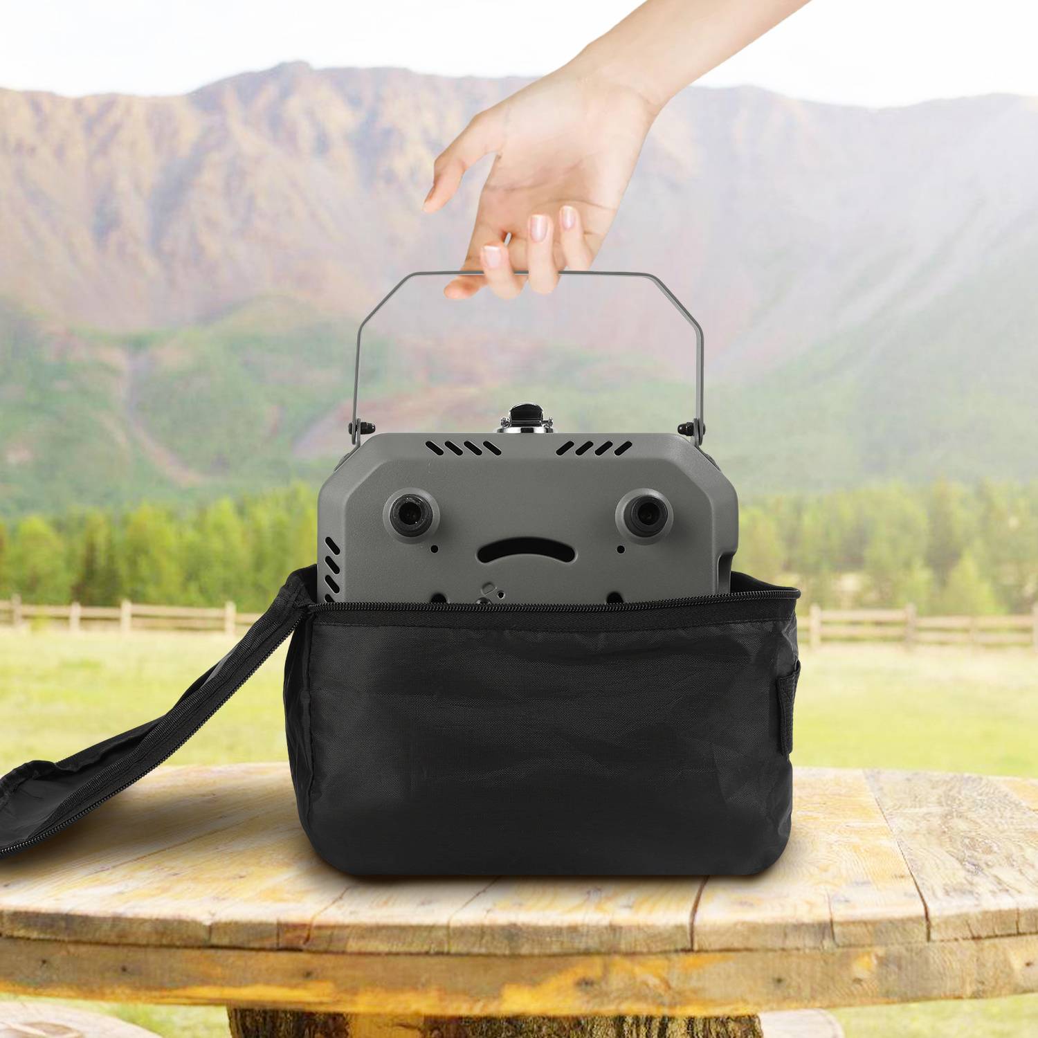Eine Hand legt einen Projektor in eine schwarze Transporttasche auf einem Holztisch, mit einer malerischen Berglandschaft im Hintergrund.