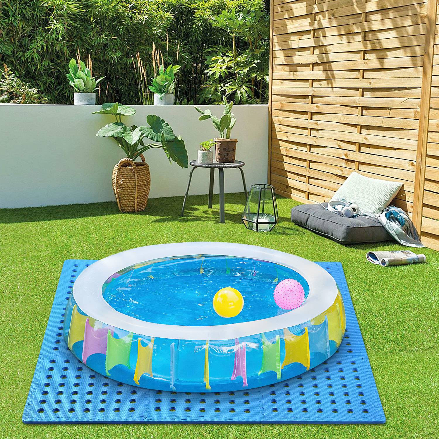 Ein aufblasbares Kinderbecken auf Gras mit gestreiftem Design, gefüllt mit Wasser und zwei farbenfrohen Bällen, umgeben von Gartenpflanzen und Sitzgelegenheiten.