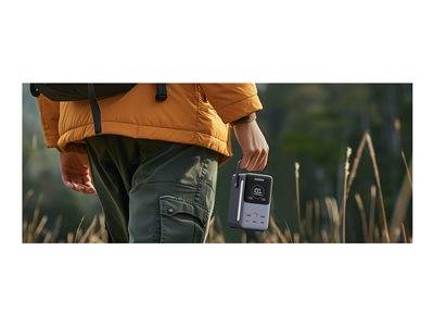 Eine Person in einer orangefarbenen Jacke und grüner Hose geht über ein Feld und hält einen tragbaren Vintage-Kassettenrekorder in der Hand.