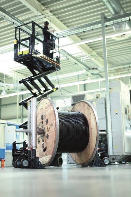 Eine große Spule schwarzes Kabel steht in einer Industrieumgebung. Oben bedient eine Person einen Scherenhubwagen, um Ausrüstung zu inspizieren.