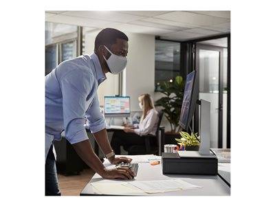 Ein Mann mit einer Maske arbeitet an einem Computer in einem Büro, während eine Frau im Hintergrund ebenfalls einen Computer benutzt.