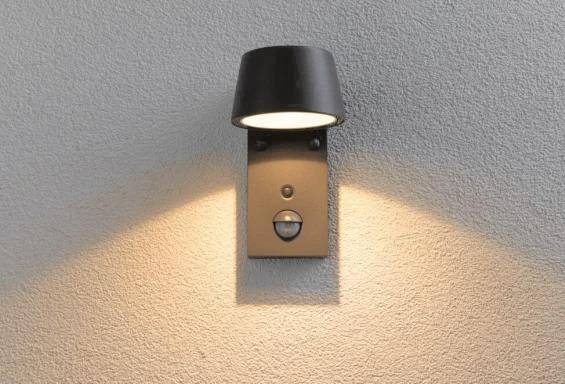 Eine an der Wand montierte schwarze Außenleuchte mit Bewegungsmelder, die eine strukturierte Wand mit nach unten gerichtetem Licht beleuchtet.