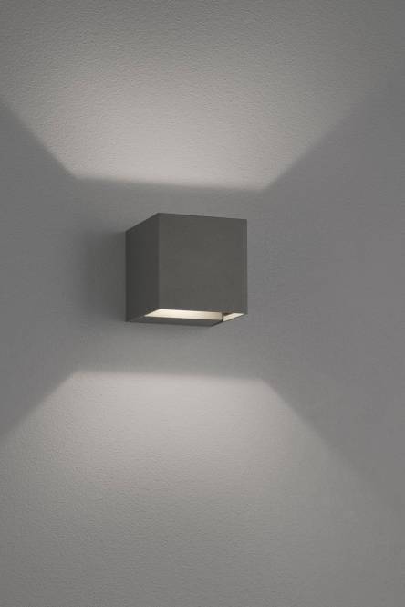 Ein modernes quadratisches Wandleuchten-Modell strahlt weiches Licht nach oben und unten auf eine graue Wand ab und schafft eine minimalistische und elegante Atmosphäre.