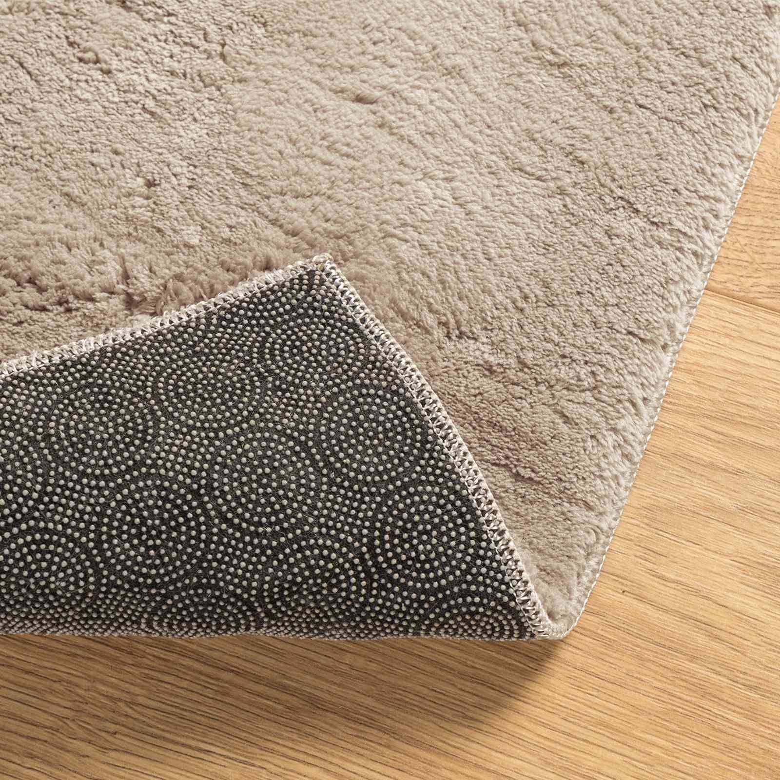 Ein rechteckiger beiger Teppich mit einer rutschfesten gepunkteten Rückseite, der teilweise umgeklappt ist und auf einem Holzboden liegt, zeigt Textur- und Designdetails.