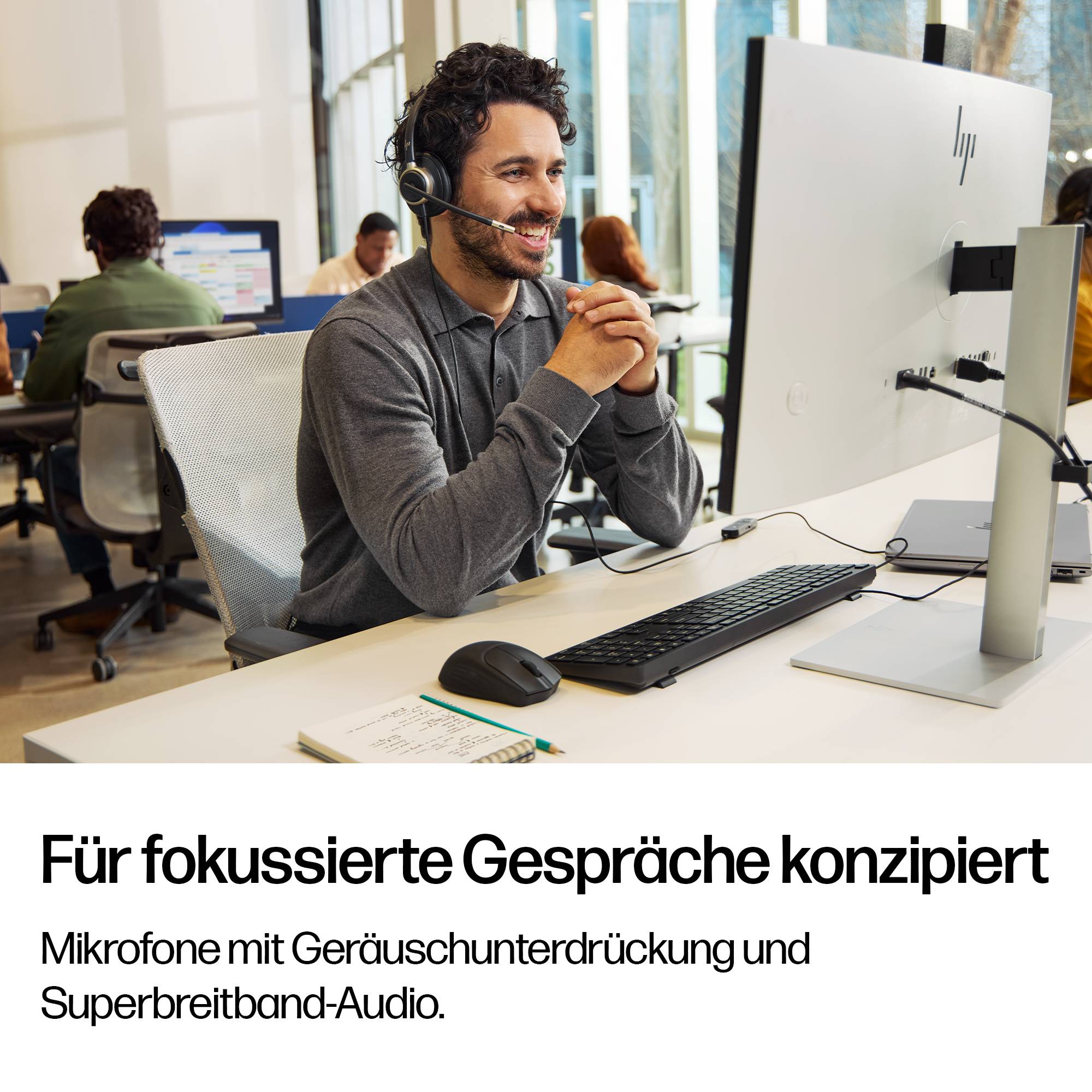 Ein Kundendienstmitarbeiter mit Headset lächelt auf einen Desktop-Monitor in einem modernen Büro; der Anzeigentext besagt, dass das Mikrofon für konzentrierte Gespräche mit Geräuschunterdrückung und Super-Breitband-Audio ausgelegt ist.