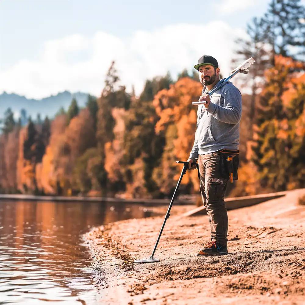 Mann, der mit einem Metalldetektor an einem sandigen Seeufer steht, eine Schaufel hält, neben ruhigem Wasser und Herbstbäumen.