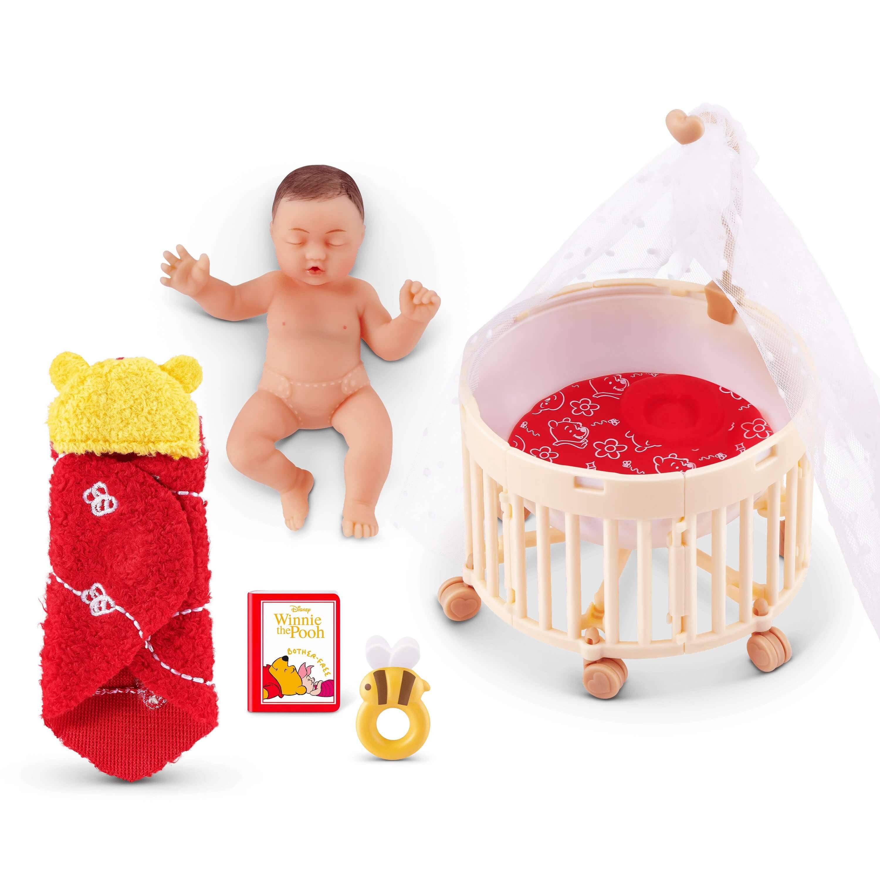 Eine Puppe liegt neben einem Babybett, in eine rote Decke mit gelbem Bärenkopf-Design gewickelt, neben einem kleinen Buch und einem bienenförmigen Spielzeug.