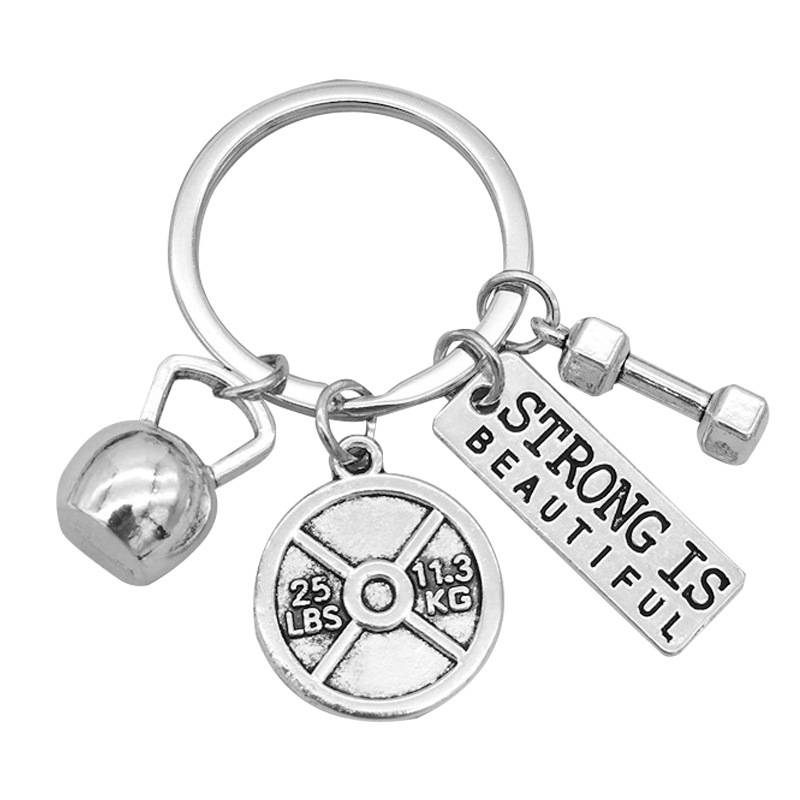 Schlüsselanhänger mit Anhängern einer Kettlebell, einer Hantelscheibe mit der Aufschrift 25 lbs 11,3 kg und einem Etikett mit der Aufschrift „Strong is Beautiful