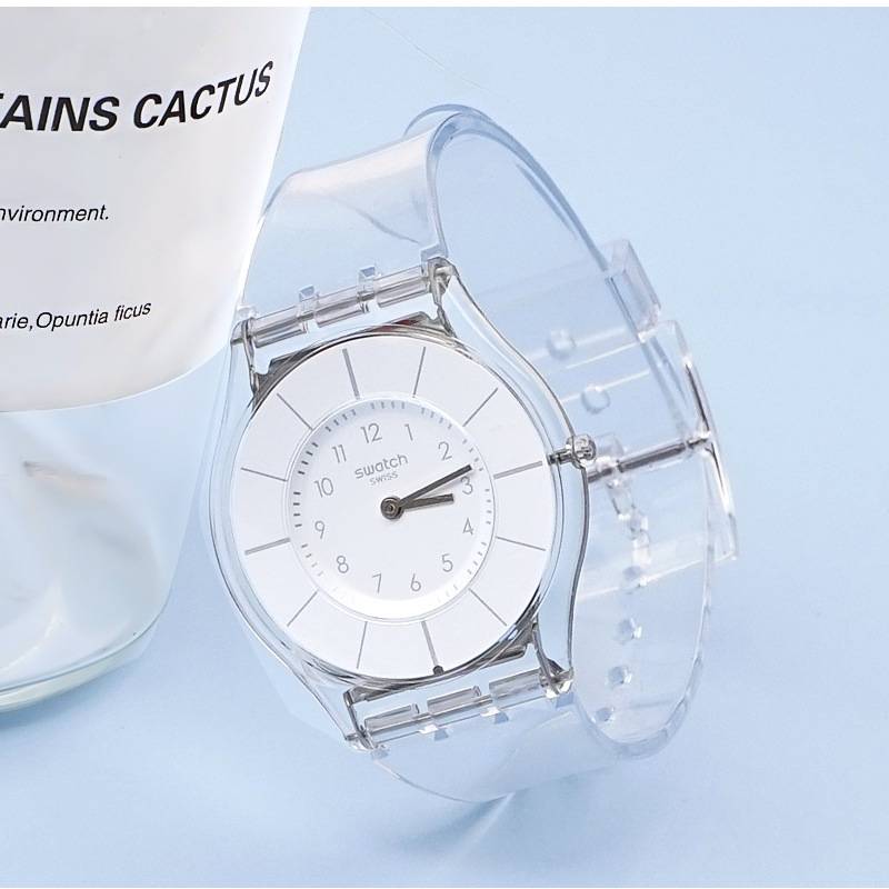 Eine klare Armbanduhr mit weißem Zifferblatt und silbernen Zeigern, neben einem teilweise sichtbaren Produkt mit der Aufschrift „CACTUS". Hellblauer Hintergrund.