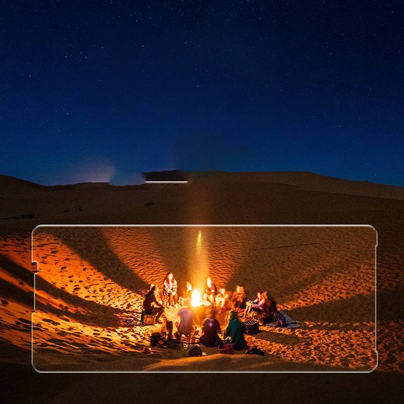 Eine Gruppe von Menschen sitzt um ein leuchtendes Lagerfeuer in einer Wüste bei Nacht, wobei das Licht Schatten auf die sie umgebenden Sanddünen wirft.