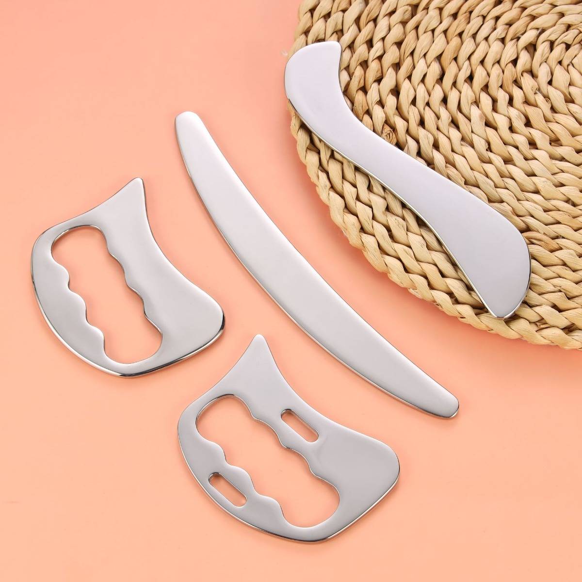 Vier Gua-Sha-Werkzeuge aus Edelstahl auf einer geflochtenen Matte und pfirsichfarbenem Hintergrund, die verschiedene Formen zur Muskelentspannung und Massage zeigen.