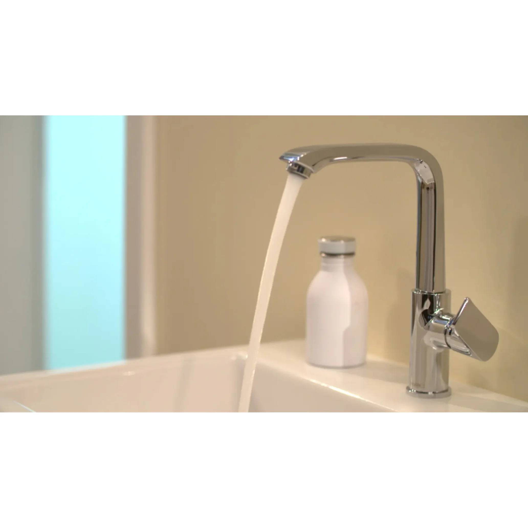 Wasser, das aus einem verchromten Badezimmerhahn in ein Waschbecken fließt, mit einer kleinen weißen Flasche dahinter.