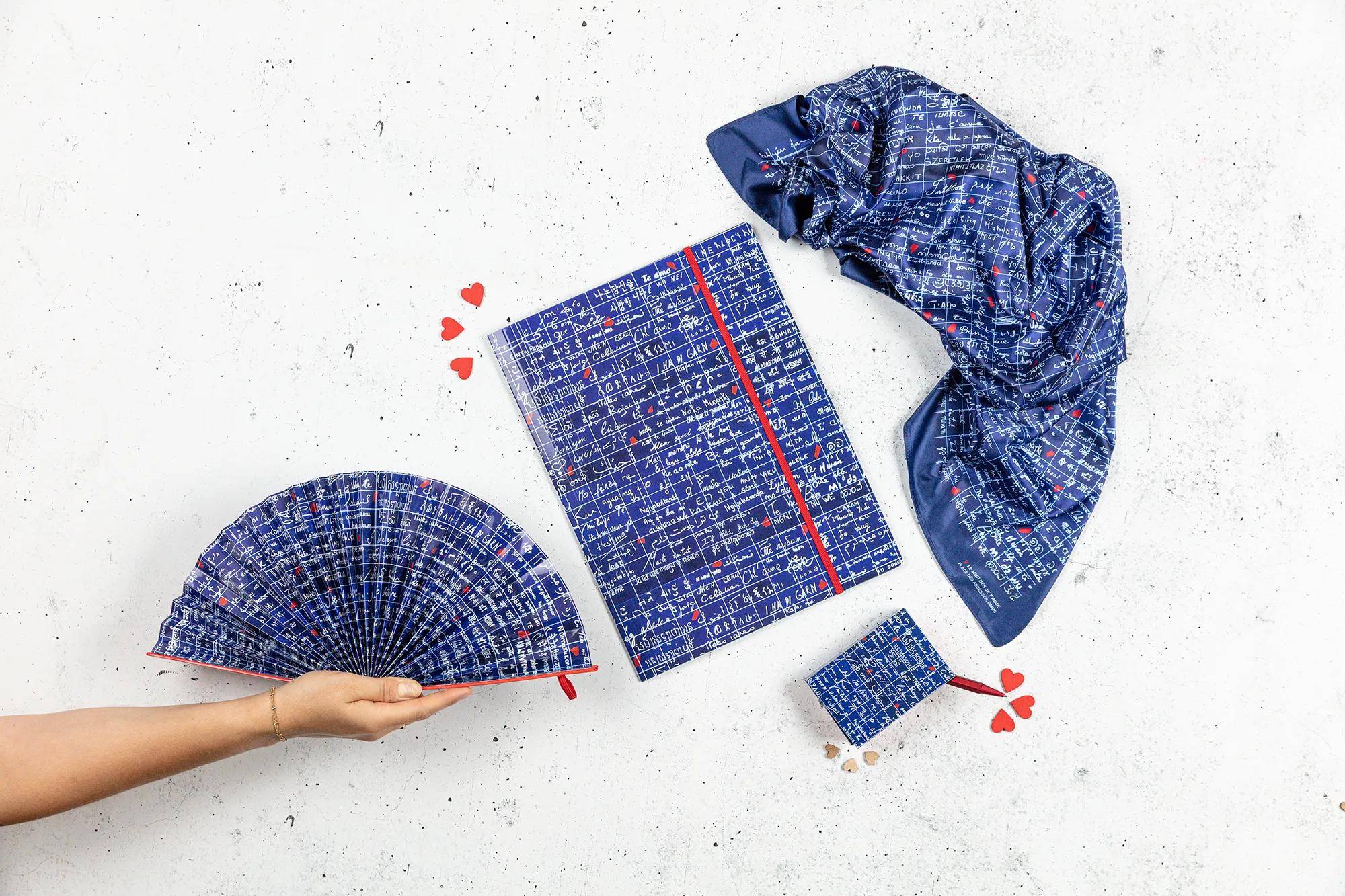 Hand hält blauen gemusterten Faltfächer neben einem passenden quadratischen Tuch, Schal und kleinem Beutel auf weißem Hintergrund mit roten Herzakzenten.