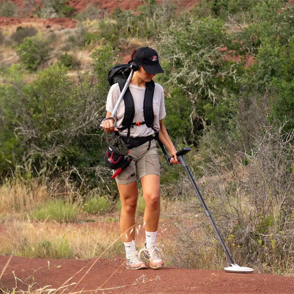Frau beim Wandern auf einem roten Schotterweg mit Rucksack und Wanderstock in einer strauchigen, felsigen Landschaft
