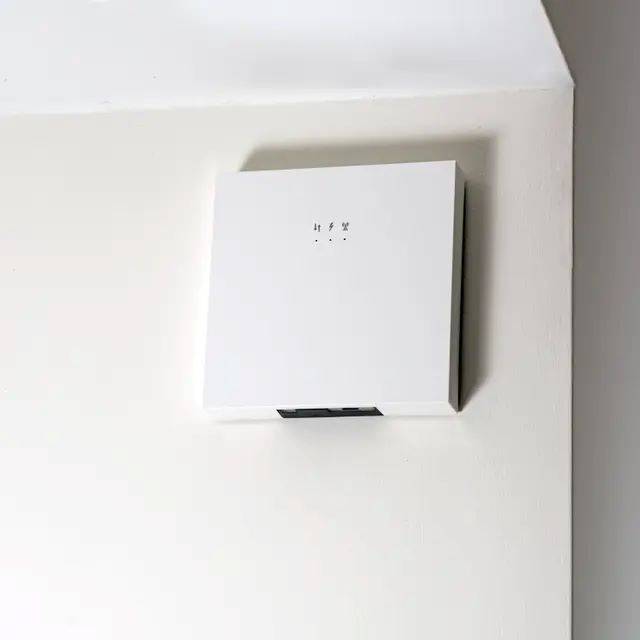 Ein weiß angebrachtes quadratisches elektrisches Bedienfeld oder Schaltkasten an einer ebenen Innenwand mit einer kleinen schwarzen Öffnung am unteren Ende.